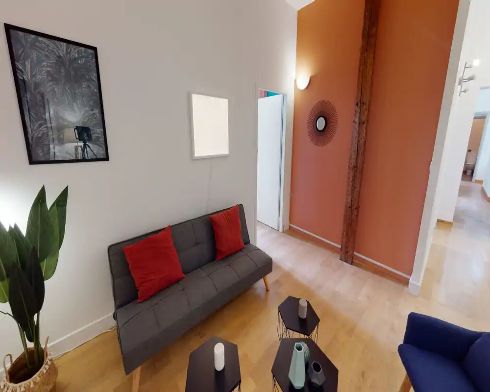 Salon moderne et lumineux avec un canapé gris compact et coussins rouges, tables basses décoratives et mur d'accent terre cuite; idéal pour présenter un coin détente chaleureux.