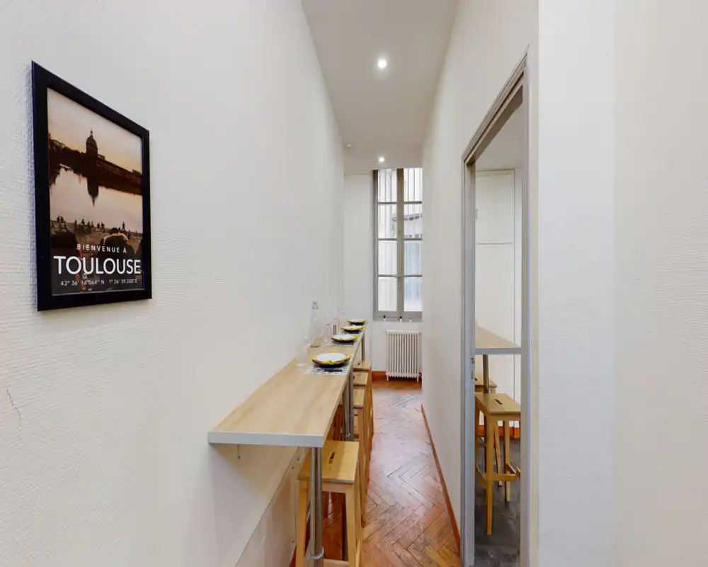 Espace repas étroit avec un long plan en bois et des tabourets dressés pour les repas ; lumière de la fenêtre et parquet en chevrons donnent une atmosphère chaleureuse et compacte.