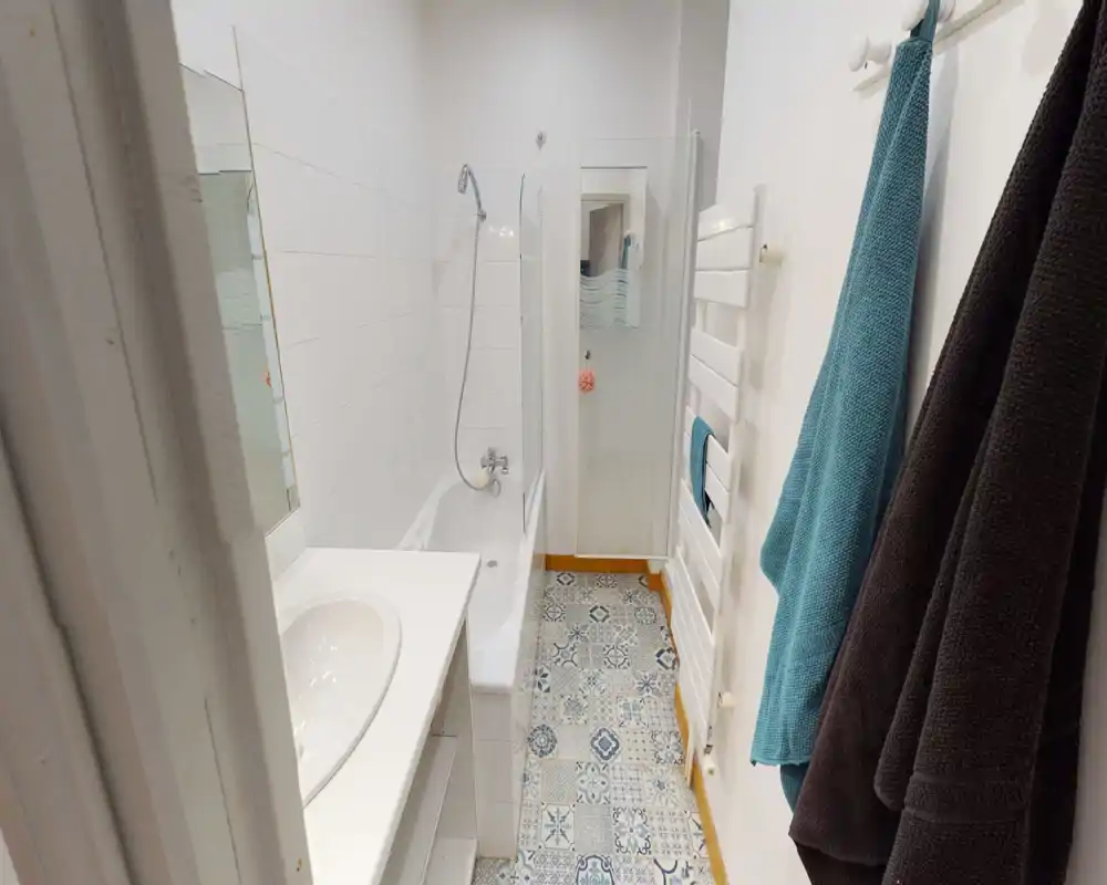 Salle de bain compacte et lumineuse avec baignoire et douche murale, carrelage sol à motifs et porte-serviettes — propre et fonctionnelle.