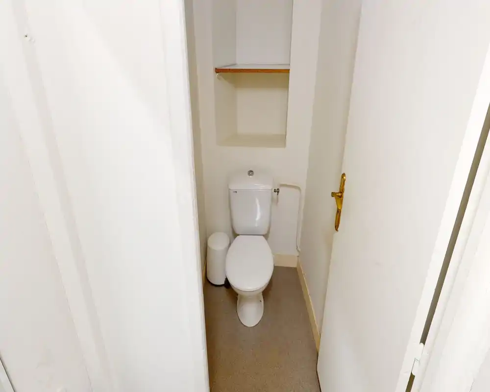 Petite pièce WC avec toilette blanche à réservoir, petite poubelle et niche étagère intégrée au-dessus. Murs neutres et sol pratique pour un WC invité fonctionnel.