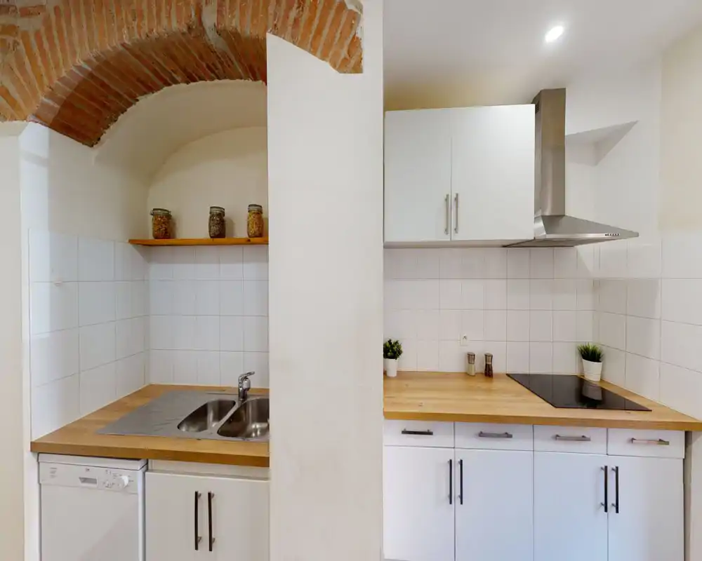 Cuisine compacte et moderne avec placards blancs, plans de travail en bois, plaque à induction et double évier. L'arche en briques apparentes et les bocaux décoratifs apportent du caractère et de la chaleur.