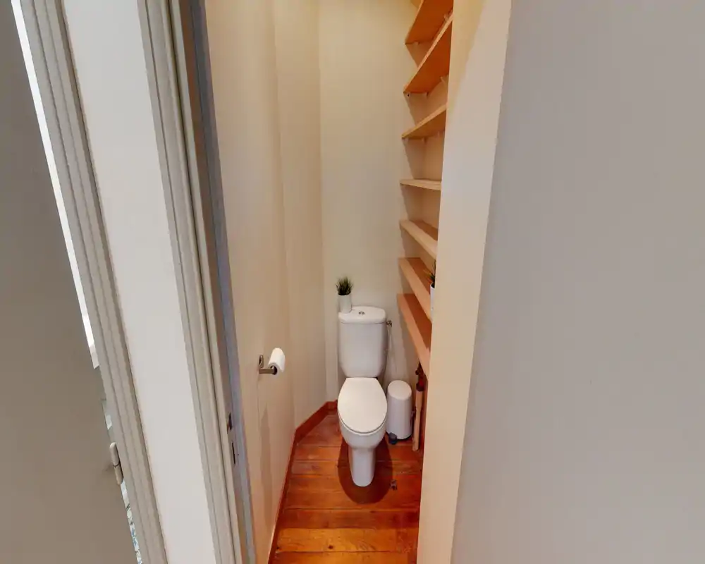 Petit WC lumineux avec toilette standard, sol en bois et étagères intégrées à droite — propre et bien éclairé.