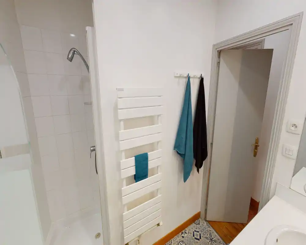 Coin salle de bain propre et bien éclairé avec douche carrelée, radiateur porte-serviettes mural et crochets avec serviettes. Murs blancs et carrelage au sol à motifs pour un rendu soigné et moderne.