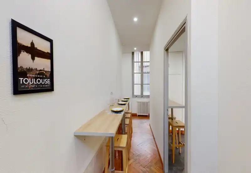 Zona pranzo stretta con bancone lungo in legno e sgabelli apparecchiati; luce dalla finestra e pavimento a spina di pesce rendono lo spazio compatto e accogliente.