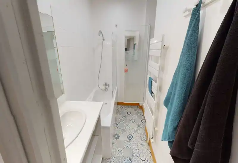 Bagno compatto e ben illuminato con vasca e doccia a muro, piastrelle decorative sul pavimento e scaldasalviette — pulito e funzionale.