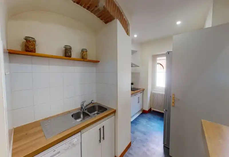 Cucina compatta con piani in legno, doppio lavello in acciaio e rivestimento a piastrelle bianche; luce naturale dalla finestra illumina l'ambiente.