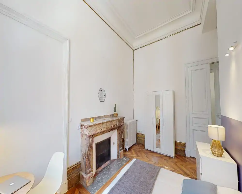Bright, elegant bedroom with a decorative marble fireplace, wooden herringbone floor and a compact double bed. The space features a wardrobe, bedside table with lamp and tasteful minimal decor — ideal for a cozy urban stay.