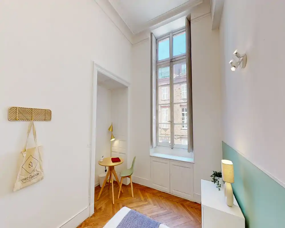 Camera da letto luminosa con soffitti alti, grande finestra, piccolo angolo studio e luce accogliente. Stile minimal pulito con pavimento in parquet caldo e tessili morbidi.