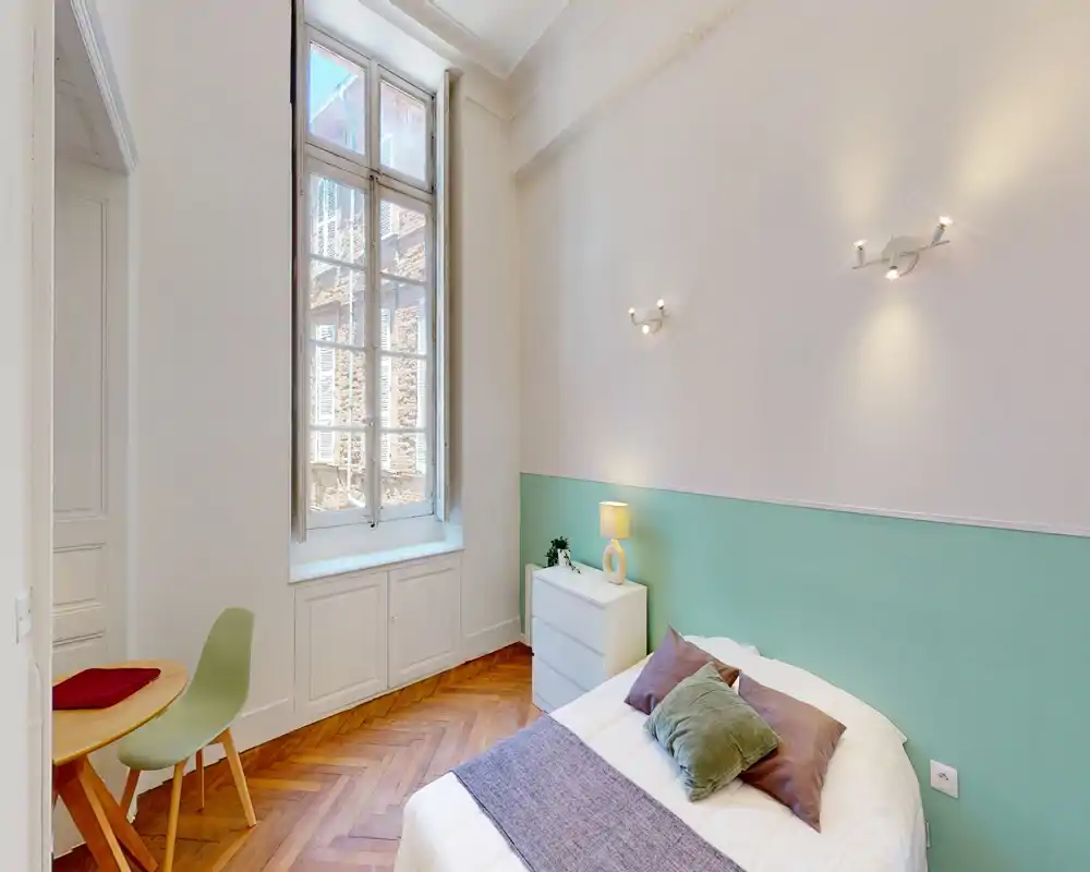 Camera da letto luminosa e accogliente con letto matrimoniale, cuscini decorativi e un comodino sotto una finestra alta che illumina la stanza con luce naturale.