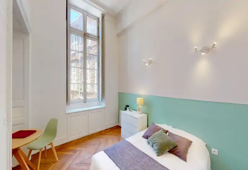 Camera da letto luminosa e accogliente con letto matrimoniale, cuscini decorativi e un comodino sotto una finestra alta che illumina la stanza con luce naturale.