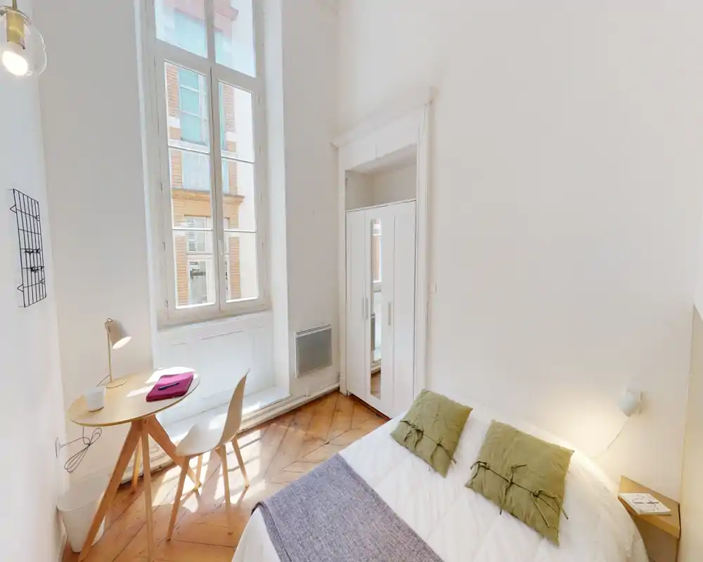 Chambre lumineuse et compacte avec un lit double confortable, grande fenêtre apportant beaucoup de lumière naturelle, petit bureau et armoire — idéale pour un séjour citadin chaleureux.