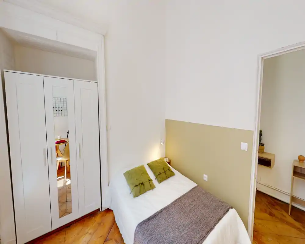 Chambre simple lumineuse et cosy avec parquet, lit bien fait avec coussins verts et armoire blanche — idéale pour un séjour confortable en ville.