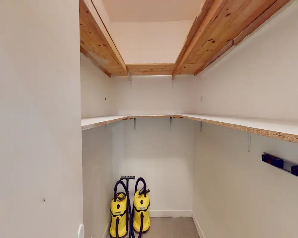 Petit local de rangement/utility avec étagères intégrées et deux aspirateurs verticaux ; espace pratique pour produits ménagers et rangement.