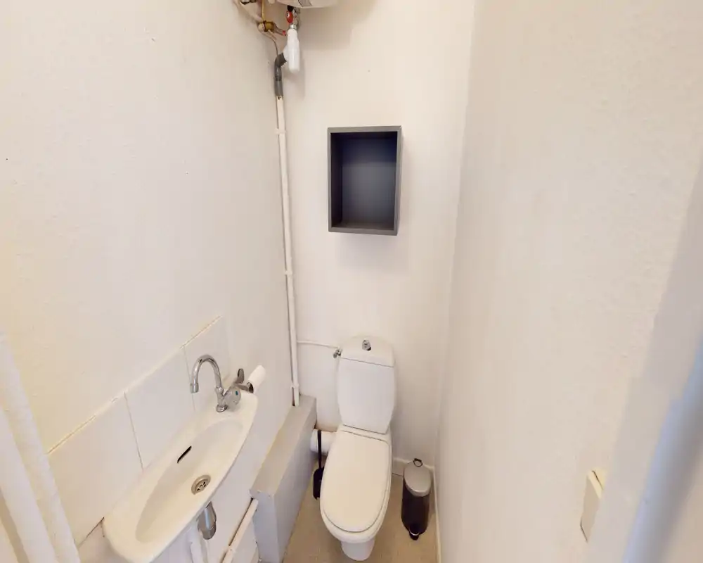Salle de bain compacte avec toilette, petit lavabo et étagère murale; murs blancs neutres et équipements pratiques, idéal pour un WC invité.