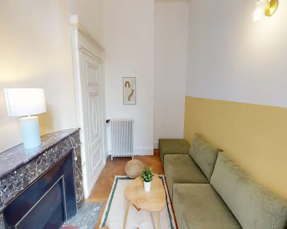 Espace salon lumineux et chaleureux avec un canapé vert olive, une petite table basse en bois et un tapis décoratif. La pièce est dotée d'une cheminée en marbre et d'un éclairage chaleureux, parfaite pour se détendre.