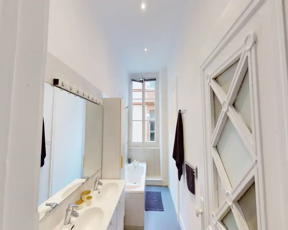 Salle de bains lumineuse avec double vasque, grand miroir, baignoire près d'une haute fenêtre et porte-serviettes bien rangés—parfaite pour un séjour confortable.