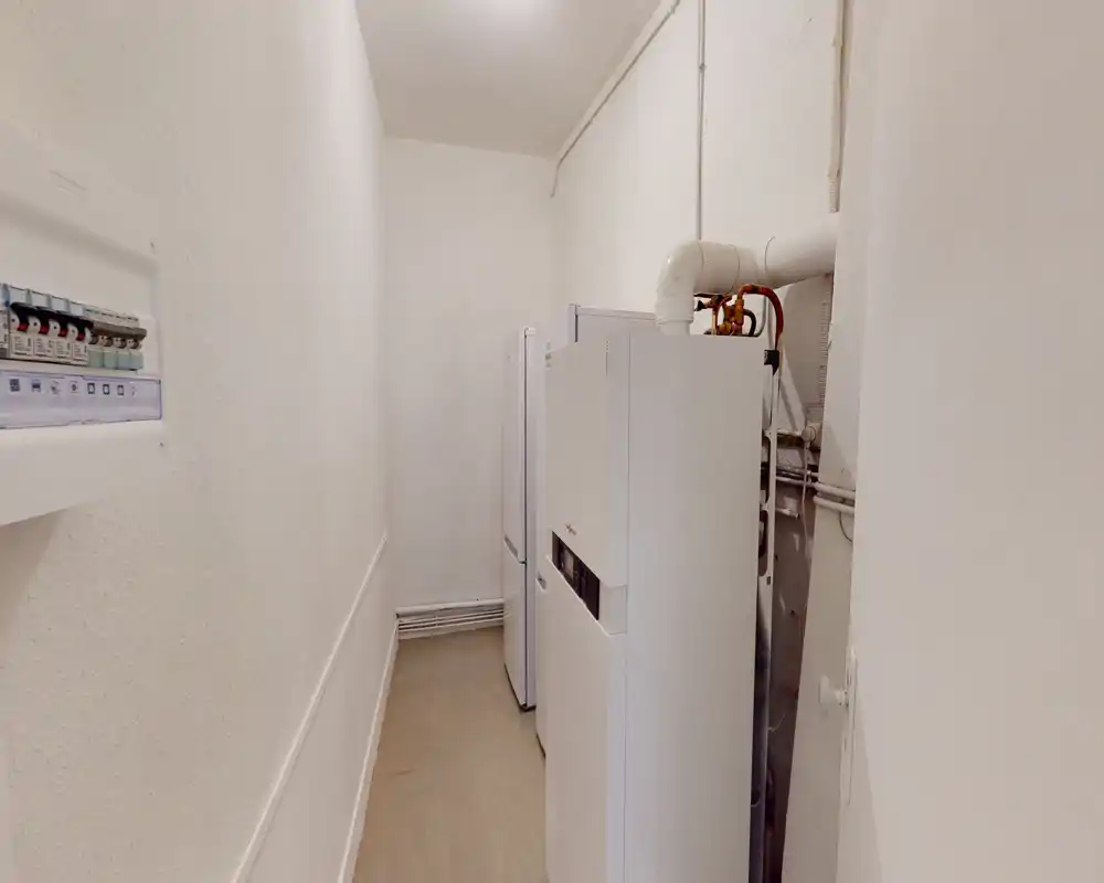 Couloir technique/locale technique étroit avec chaudières et tableau électrique ; espace de service fonctionnel aux murs blancs et à l'aménagement pratique.