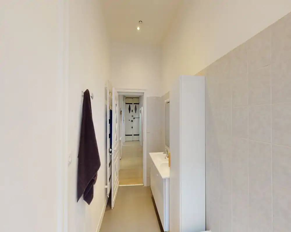 Salle de bain étroite et moderne avec lavabo et colonne de rangement; mur carrelé d’un côté et porte-serviette près de la porte. La vue vers le couloir montre l’agencement et les finitions.