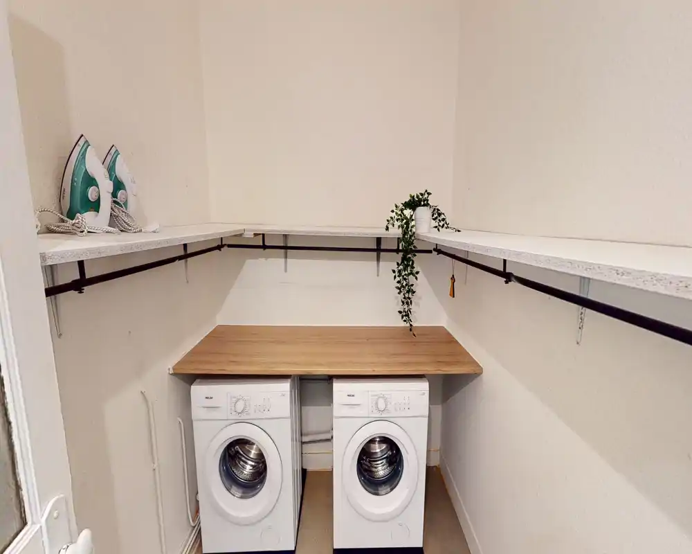 Buanderie/espace utilitaire aménagé avec deux lave-linge à hublot, un plan de travail en bois et des étagères. Lumineux, propre et pratique pour les tâches ménagères.