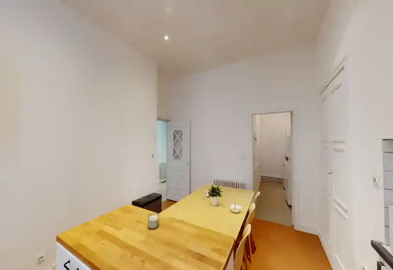 Zona pranzo luminosa e semplice con tavolo in legno, sedie e decorazioni essenziali; pareti bianche e pavimento caldo creano un ambiente pulito e accogliente.