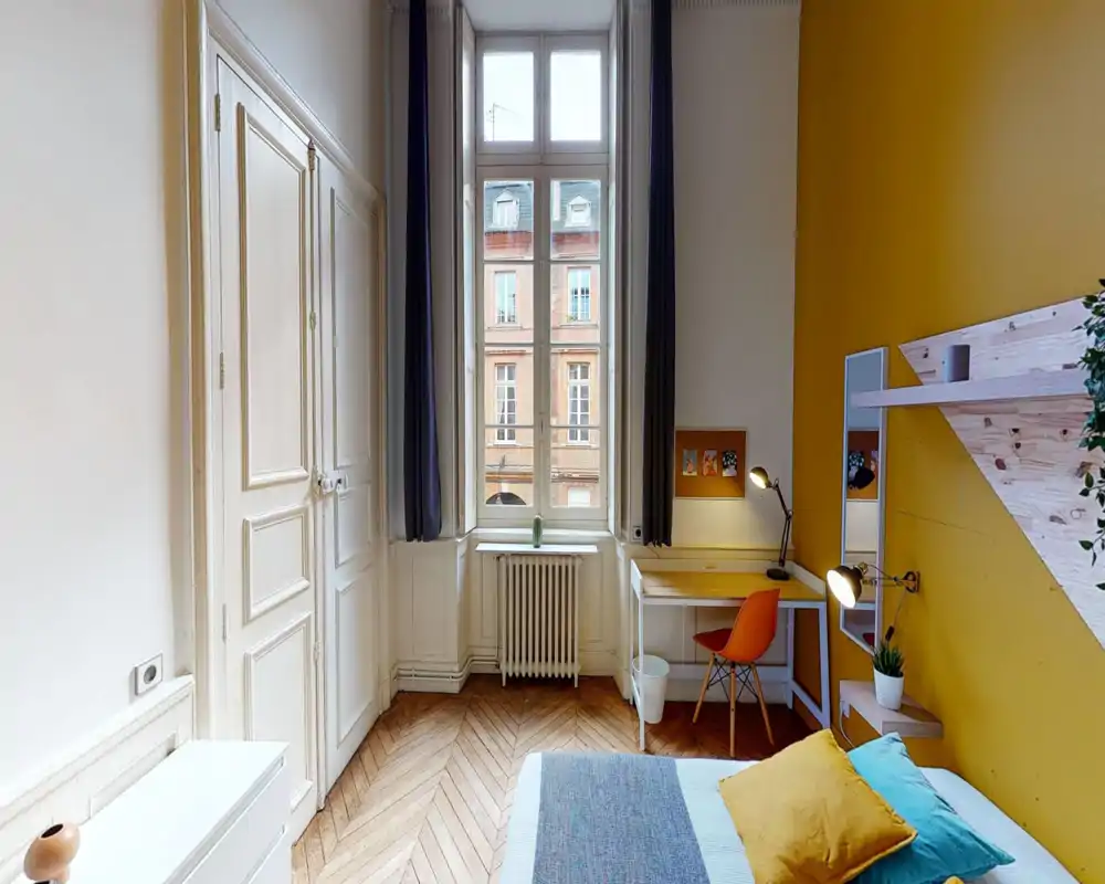 Bright, stylish bedroom with a large central window, wooden herringbone floor, cozy pillows and a functional desk area — ideal for both rest and work.
