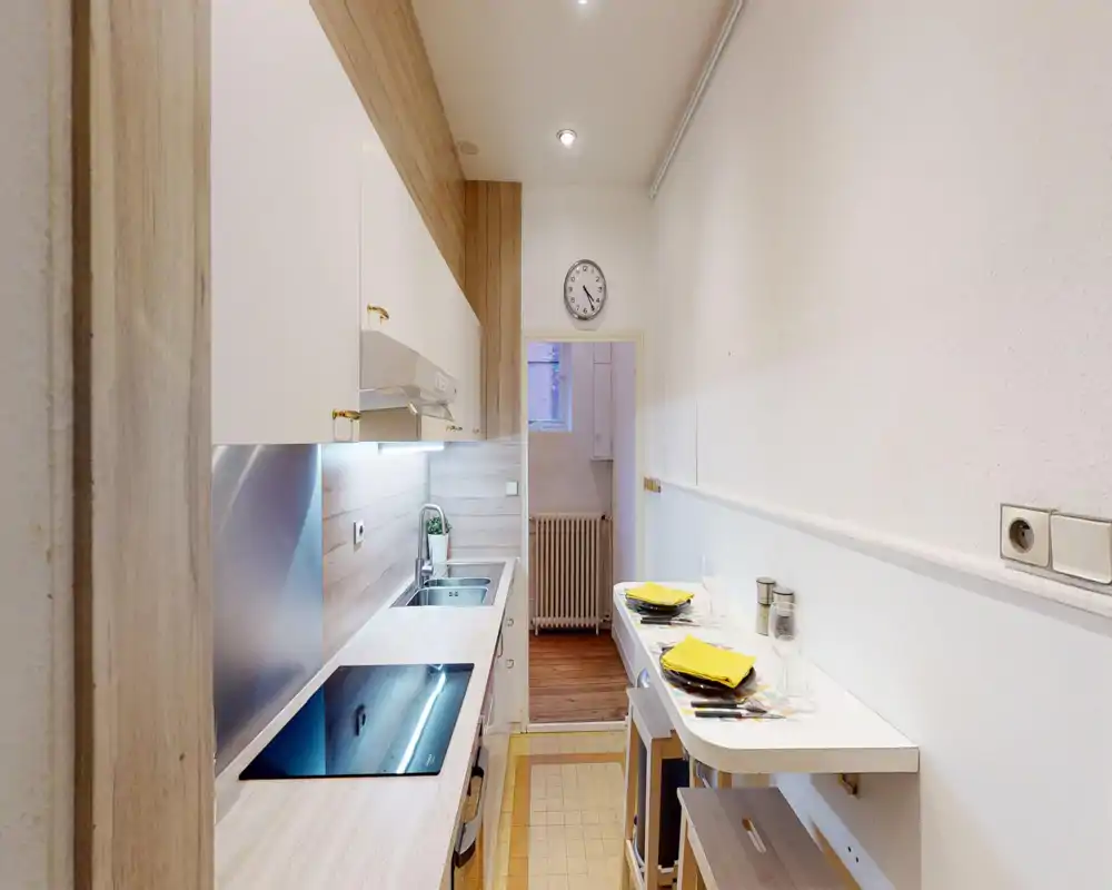 Compact, modern kitchen with a built-in induction hob, stainless sink and white cabinets; a small breakfast bar set with placemats and glasses creates a practical dining spot.