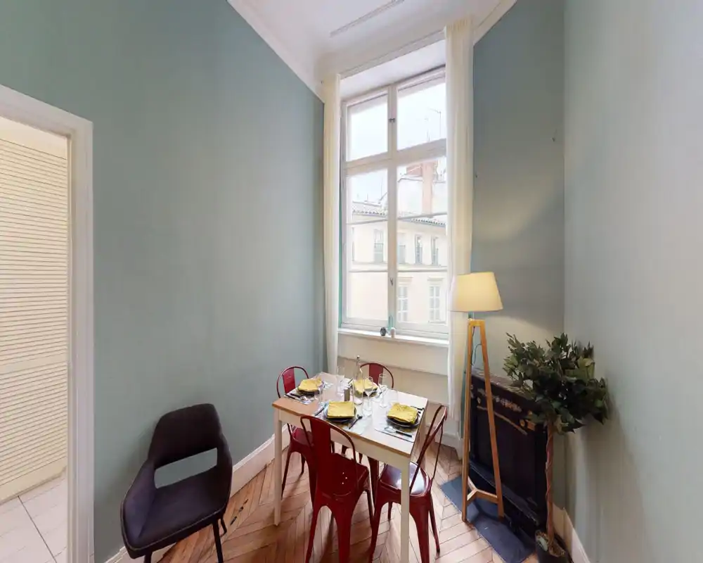 Bright, styled dining area with a window view. A compact table set for four with red metal chairs, a floor lamp and indoor plant create a cozy mealtime corner.
