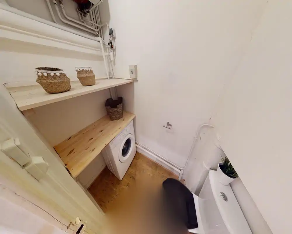 Compact bathroom with a visible toilet and a built-in shelf above a washing machine. Neutral white walls and wooden shelving create a simple, functional utility corner suitable for small-city apartments.