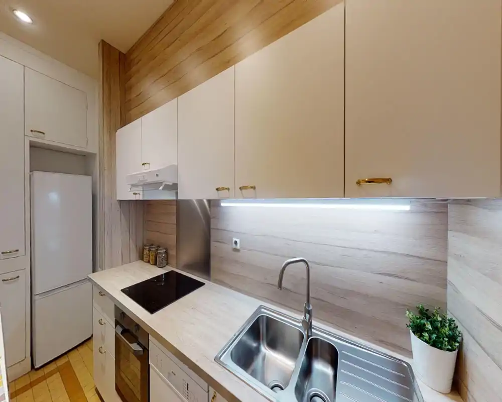 Modern compact kitchen with light wood-effect surfaces, white cabinets, stainless-steel sink and built-in appliances; under-cabinet lighting brightens the worktop and a small plant and jars add subtle decorative warmth.
