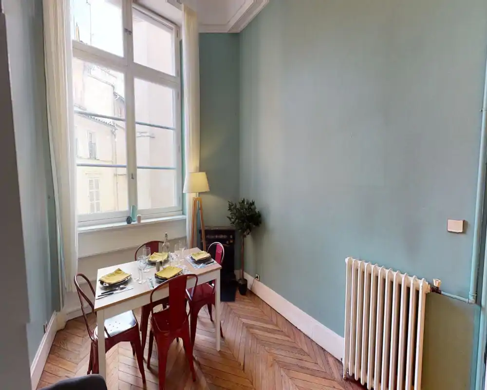 Bright dining corner with a small table set for four, red metal chairs, and a large window that fills the space with natural light.