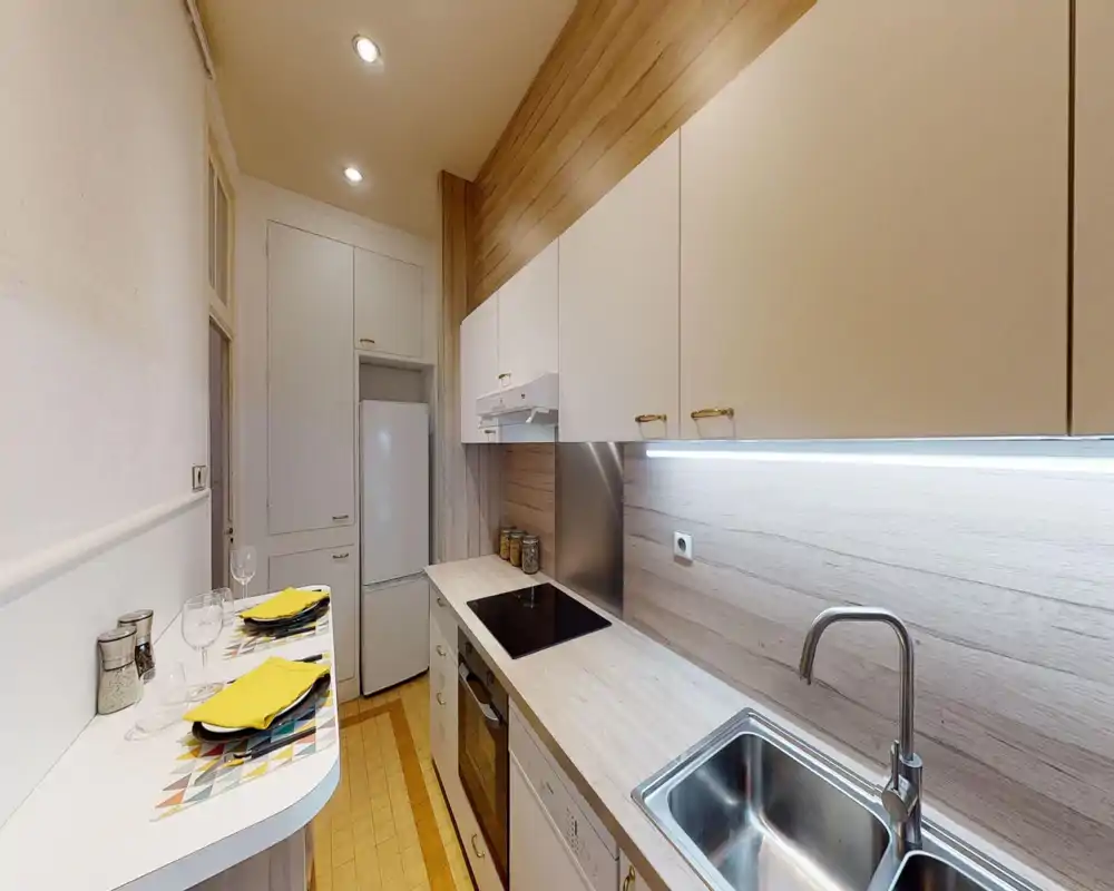 Modern galley kitchen with light wood countertops, integrated appliances, a stainless sink and under-cabinet lighting; small breakfast bar set for two with colorful placemats.
