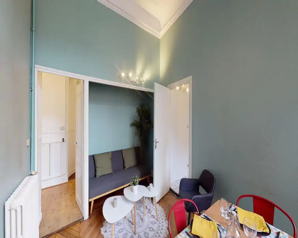 Bright and stylish living area with a modern sofa, nested coffee tables and a small dining corner. Muted green walls and wood parquet create a warm, inviting atmosphere ideal for guests or relaxation.