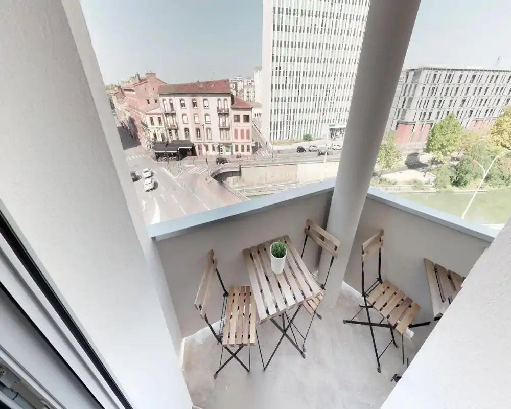 Sunny city balcony with a small wooden bistro table and folding chairs, perfect for morning coffee while overlooking a street and river view.