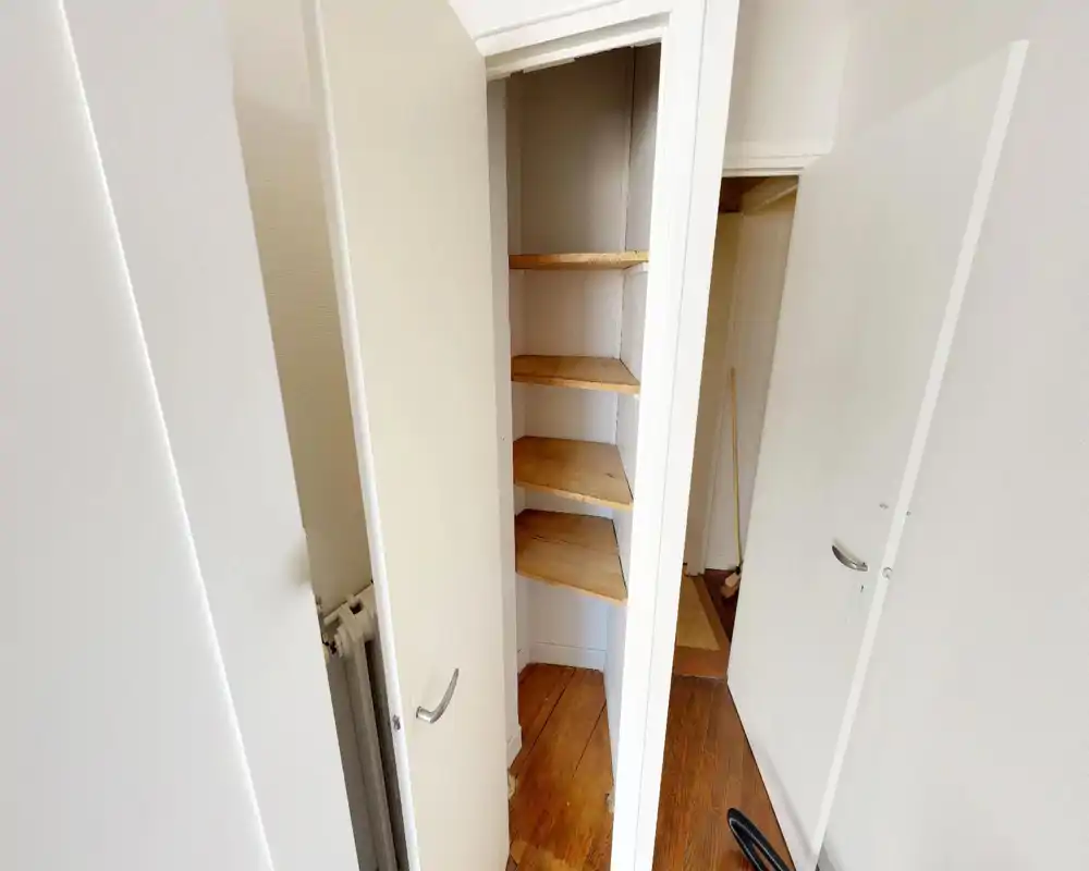 A small built-in storage/entry closet with four wooden shelves and a narrow doorway; clean white walls and wooden floor visible.