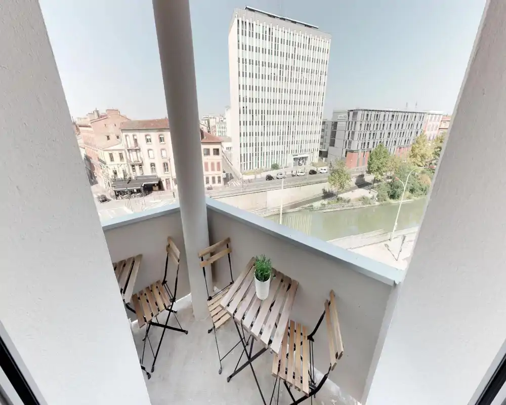 Sunny balcony with a small wooden bistro table and four folding chairs, overlooking a canal and city buildings — ideal for morning coffee or a quiet evening.