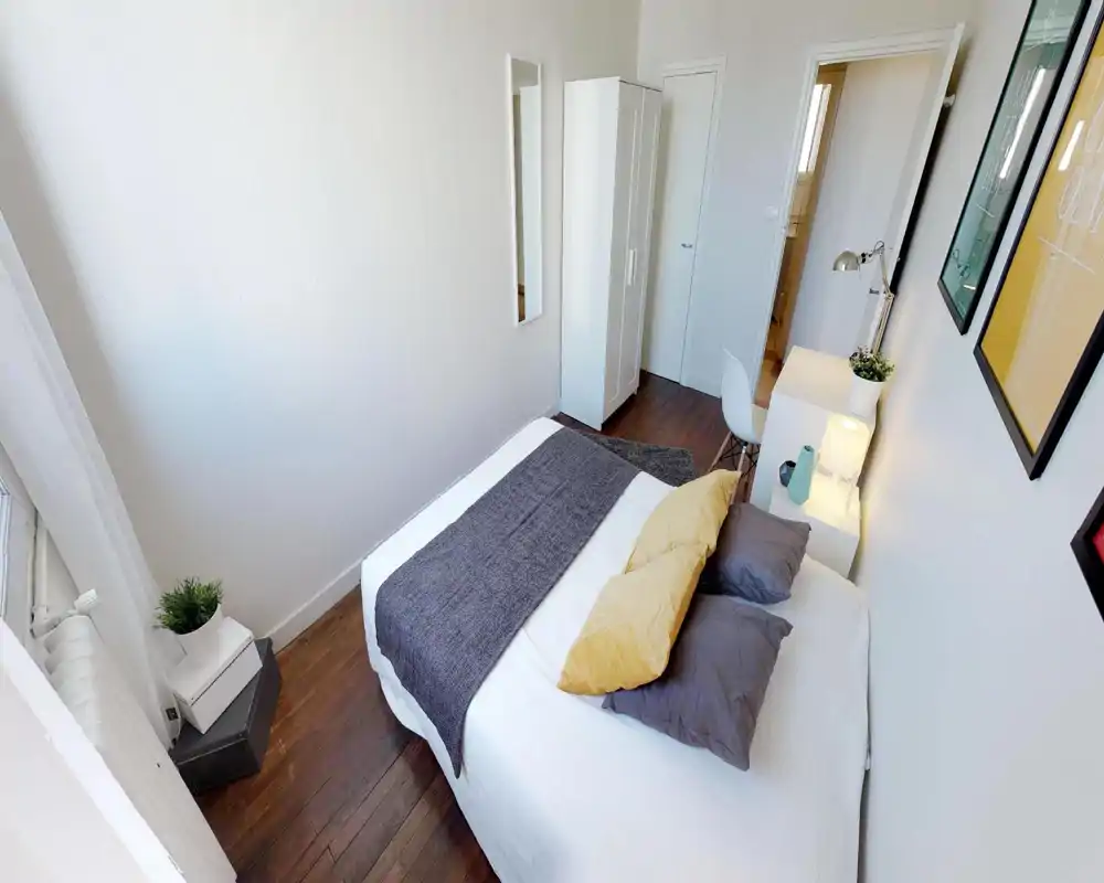 Dormitorio luminoso y ordenado con cama doble bien arreglada, cojines decorativos y mesita con lámpara; paredes blancas y suelo de madera aportan un estilo moderno.