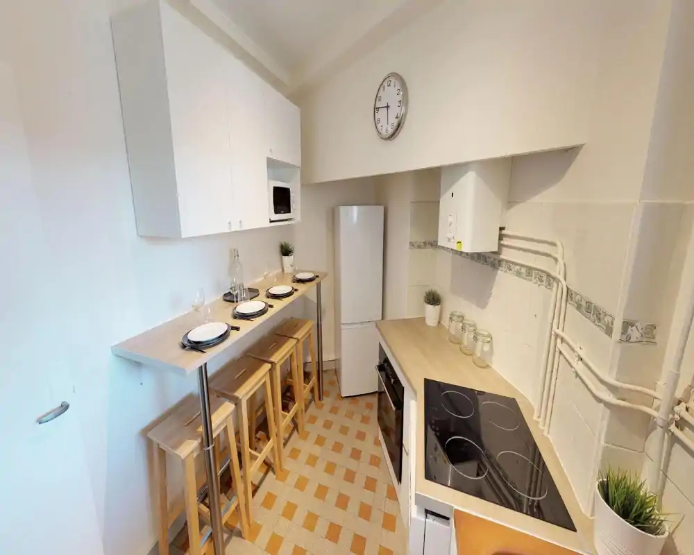Compact, well-equipped kitchen with a breakfast bar, four wooden stools, electric hob and a tall fridge. Light wood countertops, white cabinets and small plants create a neat, welcoming space ideal for casual meals.