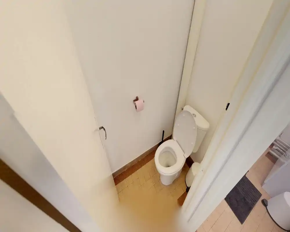 Compact bathroom with a visible toilet, open seat, toilet brush and a small waste bin; pale walls and tiled floor provide a simple, functional space.