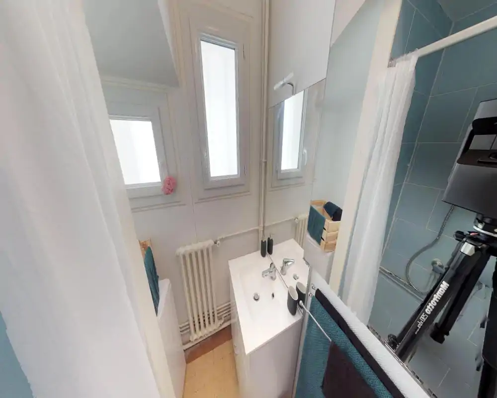 Bright compact bathroom with a white sink, mirror, radiator and a shower with blue tiles. Clean, well-lit space suitable for short-term rentals.