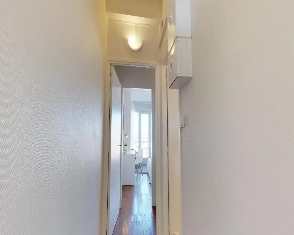Narrow, well-lit entrance corridor leading to a bright room. Clean white walls and wooden floor create a simple, welcoming first impression.