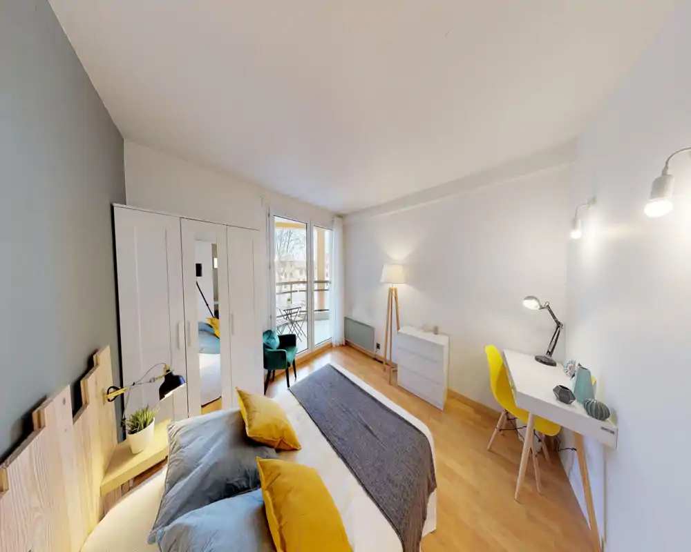 Bright, modern bedroom with a comfortable double bed, decorative cushions and a small workspace. Large balcony doors provide natural light and a cozy reading chair near the window.