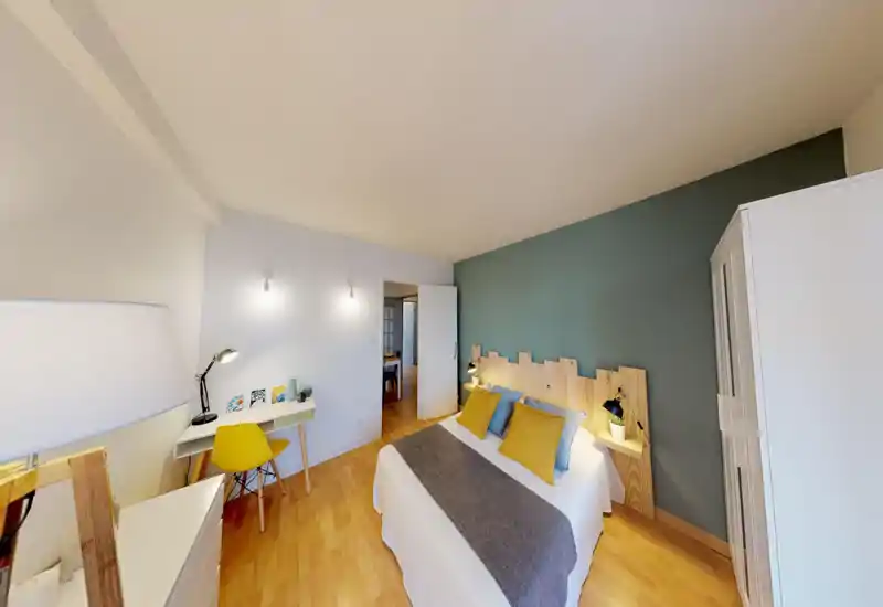 Bright, stylish bedroom with a double bed against a feature teal wall, wooden headboard and coordinated yellow and blue cushions. A small desk and chair create a practical workspace while warm wooden floors and bedside lamps add a cozy atmosphere.