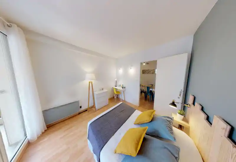 Bright, modern bedroom with a large bed dressed in blue and yellow cushions, bedside lamp and a minimalist desk area. Large window with sheer curtains provides abundant natural light and warm wooden flooring adds a cozy feel.