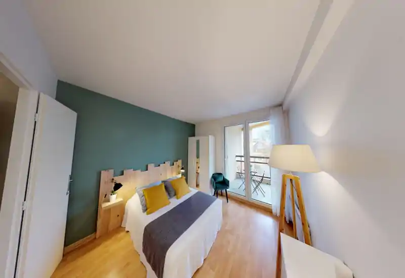 Bright, stylish bedroom with a double bed, wooden headboard and balcony access. The room features warm wood floors, a standing lamp and decorative cushions for a cozy feel.
