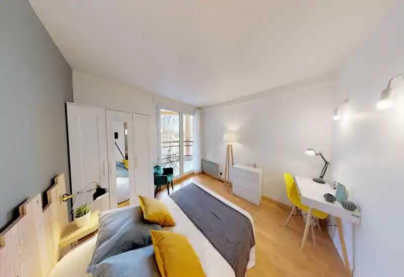 Bright, modern bedroom with a comfortable double bed, decorative cushions and a small workspace. Large balcony doors provide natural light and a cozy reading chair near the window.