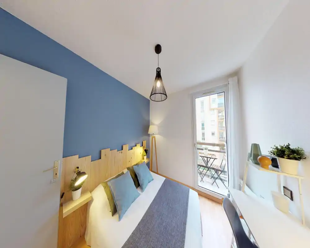 Bright, styled bedroom with a double bed, wooden headboard and soft blue accent wall. Large window/door opens to a small balcony bringing natural light and a cozy reading lamp completes the scene.