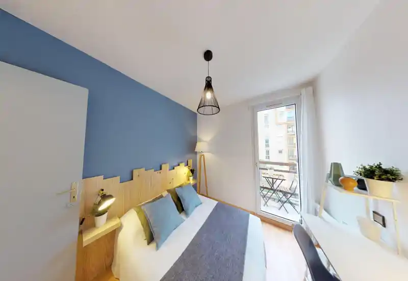 Bright, styled bedroom with a double bed, wooden headboard and soft blue accent wall. Large window/door opens to a small balcony bringing natural light and a cozy reading lamp completes the scene.