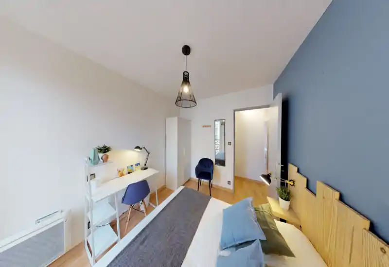 Bright, well-styled bedroom with a comfortable double bed, wooden headboard and coordinated blue accents. A small desk and chair create a practical workspace, while warm lighting and decorative cushions add a cozy feel.