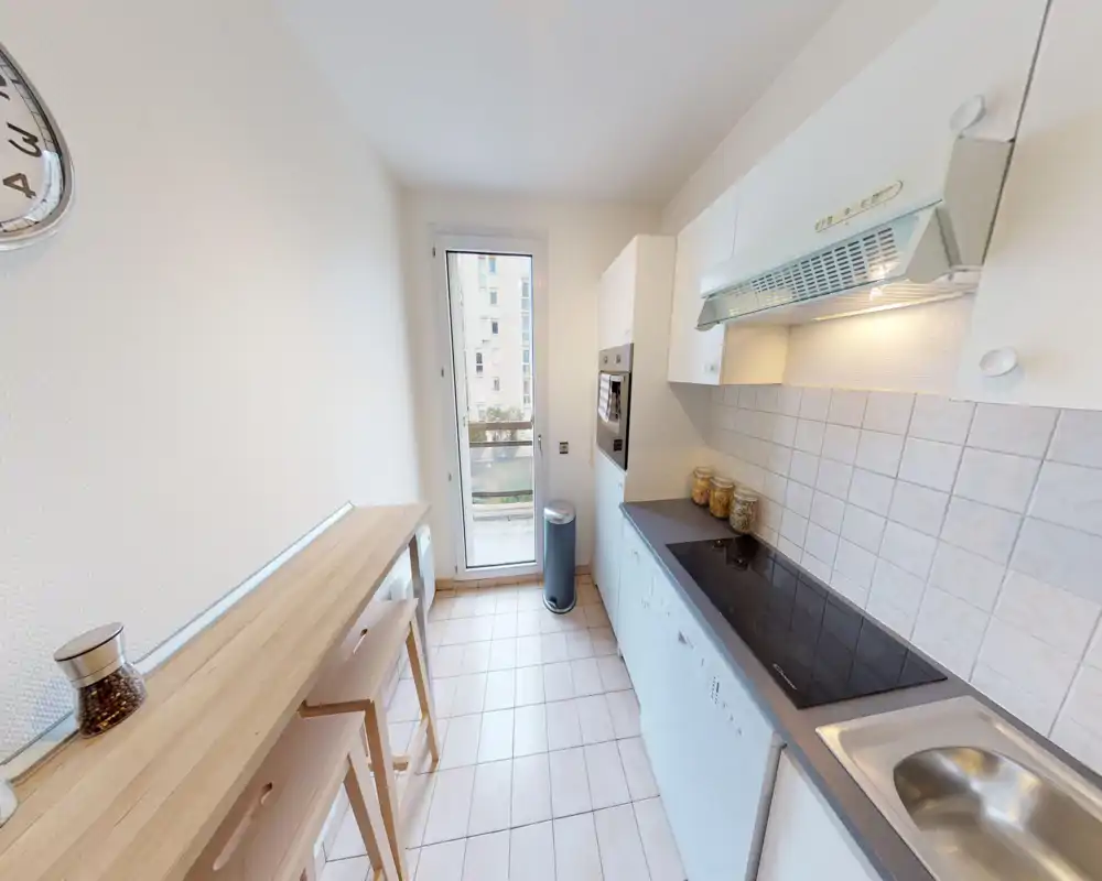 Bright, narrow kitchen with a long counter, induction hob, sink and upper cabinets. A small wooden breakfast bar with stools and glass jars give the space a warm, functional feel.