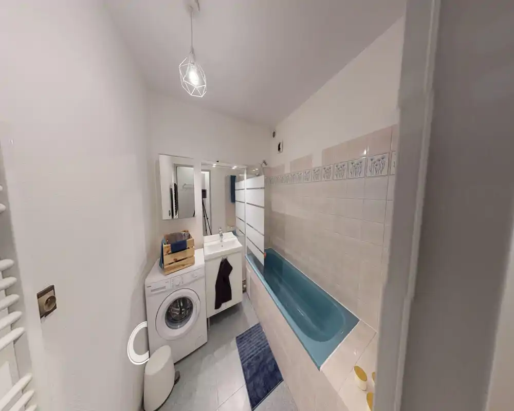 Compact, well-lit bathroom with a blue bathtub, sink with mirror, and a washing machine — clean layout ideal for a small apartment.